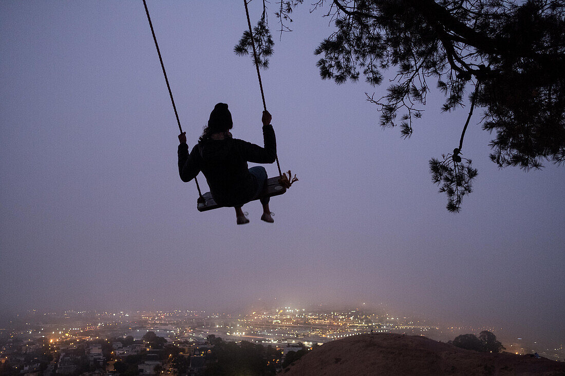 At dusk, a woman swings under a tree at Bernal Heights over San Francisco fog.; San Francisco, California, United States of America