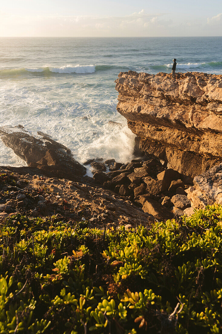 Eine Person steht auf einem zerklüfteten Bergrücken und blickt auf das Meer und die an die Küste rollenden Wellen; Praia do Guincho, Cascais, Portugal