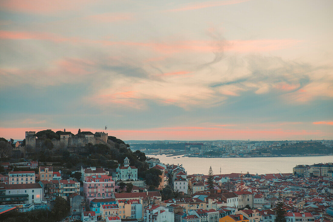 Überblick über die Altstadt von Portugals Hauptstadt Lissabon mit den pastellfarbenen Gebäuden entlang des Tejo und der St. Georgs-Burg (Castelo de Sao Jorge) auf dem Hügel im Hintergrund in der Abenddämmerung; Lissabon, Portugal
