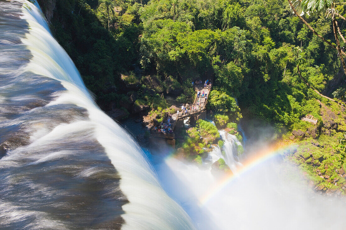 Blick über den Rand der Wasserfälle auf einen nebligen Regenbogen und Menschen auf einer Aussichtsplattform, die die Iguazu-Fälle beobachten, Iguazu Falls National Park; Puerto Iguazu, Misiones, Argentinien