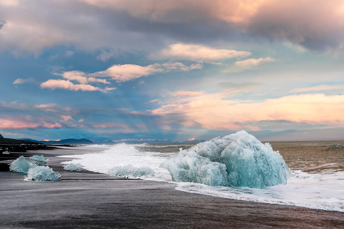 Eis aus der Jokulsarlon Gletscherlagune, das an einen schwarzen Sandstrand gespült wird, mit einer sonnenbeschienenen Bergkette am Horizont; Jokulsarlon, Vatnaj?l National Park, Island