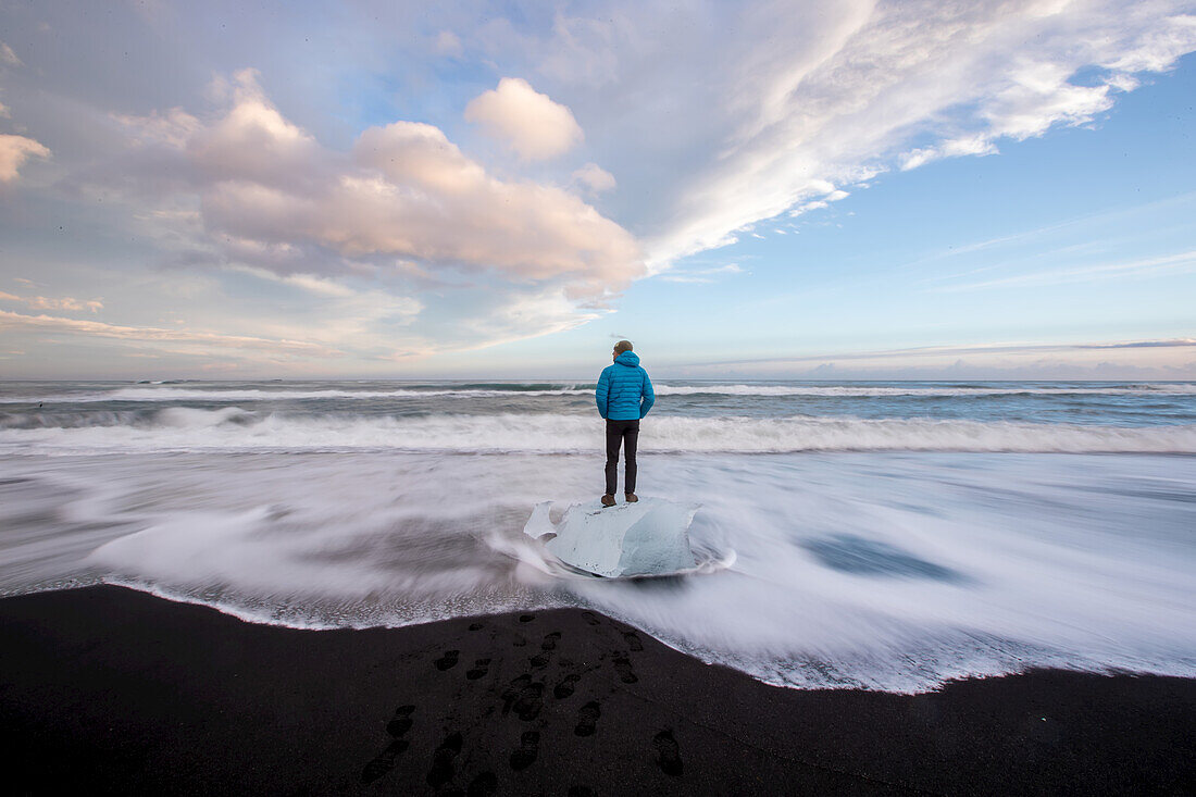 A man stands on washed up glacier ice on a black sand beach.