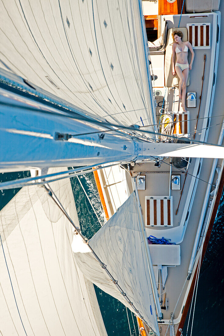 A girl sunbathes on a sailboat as seen from the tip of the mast.