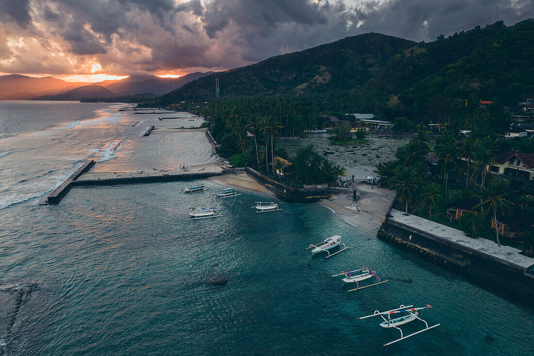 Aerial view of jukung canoes moored at a dock and along the shore of Candidasa Beach with sunlight streaming across the mountaintops under a grey cloudy sky at twilight; Candidasa Beach, East Bali, Bali, Indonesia