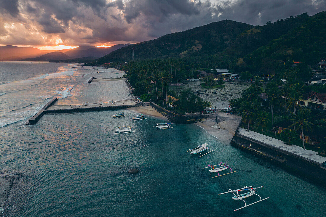 Luftaufnahme von Jukung-Kanus, die an einer Anlegestelle und am Ufer des Candidasa Beach vertäut sind, während das Sonnenlicht in der Dämmerung unter einem grauen Wolkenhimmel über die Berggipfel fällt; Candidasa Beach, Ost-Bali, Bali, Indonesien