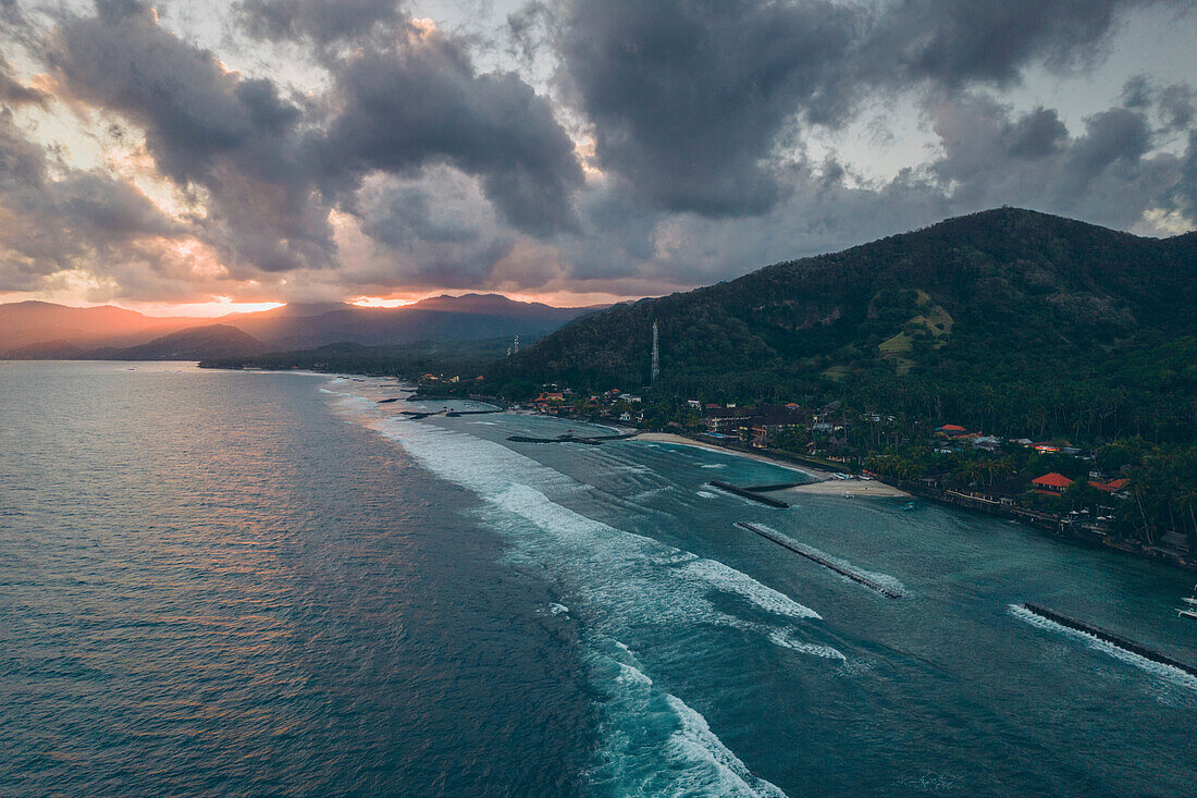 Aerial view of the docks and breakwaters along the shore of Candidasa Beach with sunlight streaming across the mountaintops under a grey cloudy sky at twilight; Candidasa Beach, East Bali, Bali, Indonesia