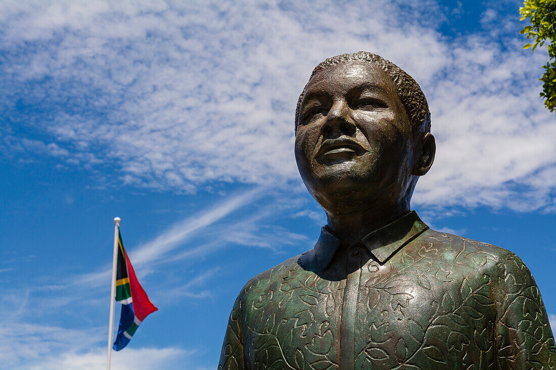 Nahaufnahme der Nelson Mandela Bronzestatue auf dem Noble Square an der Victoria and Alfred Waterfront mit der südafrikanischen Flagge vor einem bewölkten, blauen Himmel; Kapstadt, Westkap, Südafrika