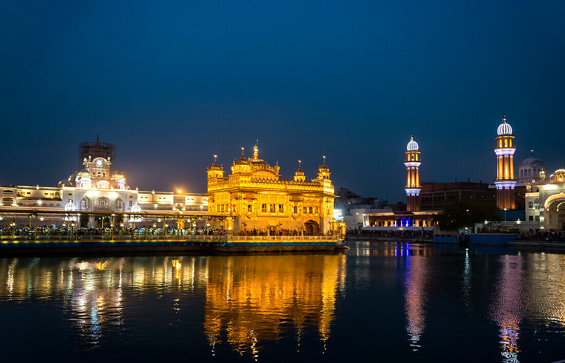Der Uhrenturm und der Eingang im gotischen Stil zum vergoldeten Goldenen Tempel (Harmandir Sahib), dem bedeutendsten heiligen Gurdwara-Komplex der Sikh-Religion mit Sarovar (Heiliger Teich) und den Türmen von Ramgarhia Bunga, die bei Nacht beleuchtet sind; Amritsar, Punjab, Indien