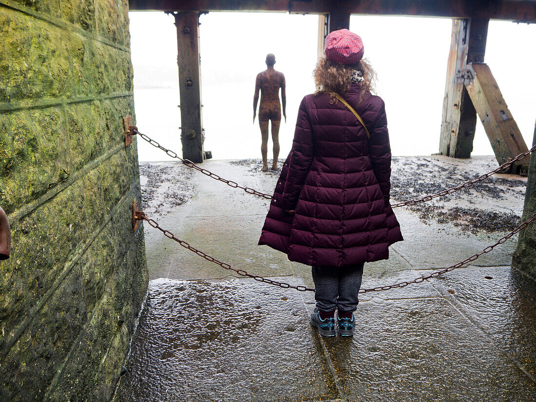Blick von hinten auf eine Frau, die als Antony Gormleys Gusseisenfigur Another Time 1999-2013 posiert und vom Folkestone Harbour Arm auf das Meer hinausschaut; Folkestone, Kent, England, Vereinigtes Königreich