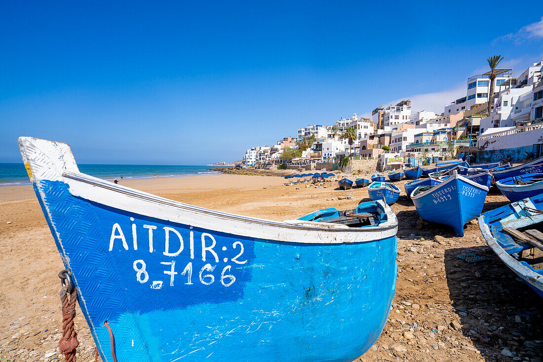 Traditionelle, leuchtend blaue Fischerboote säumen den Strand von Taghazout Village mit den weiß getünchten Gebäuden am Hang im Hintergrund; Taghazout Bay, Agadir, Marokko