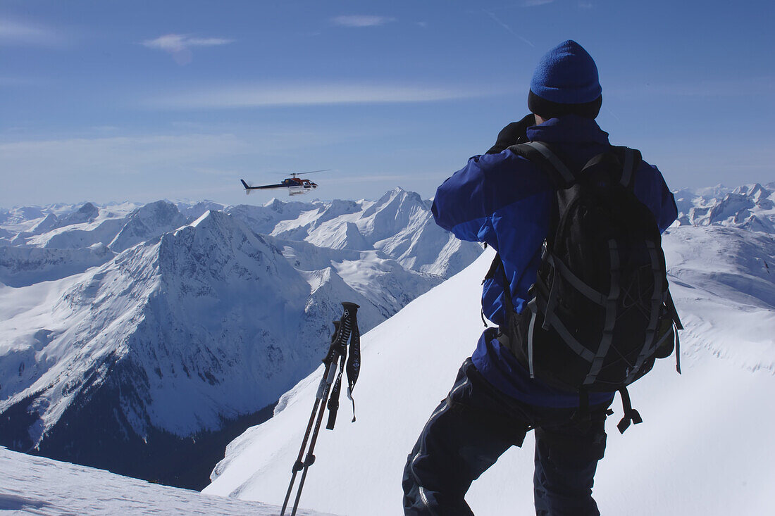 Ein Skifahrer beobachtet, wie sich ein Heli-Ski-Helikopter einem Gipfel nähert; Selkirk Mountains, British Columbia, Kanada.