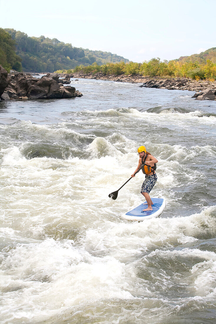 A man on a stand up paddle board runs rapids in the Potomac River.; Bethesda, Maryland.