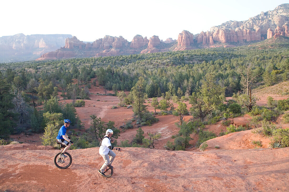 Two men on unicycles ride a rocky ridge in the Arizona desert.; Sedona, Arizona.