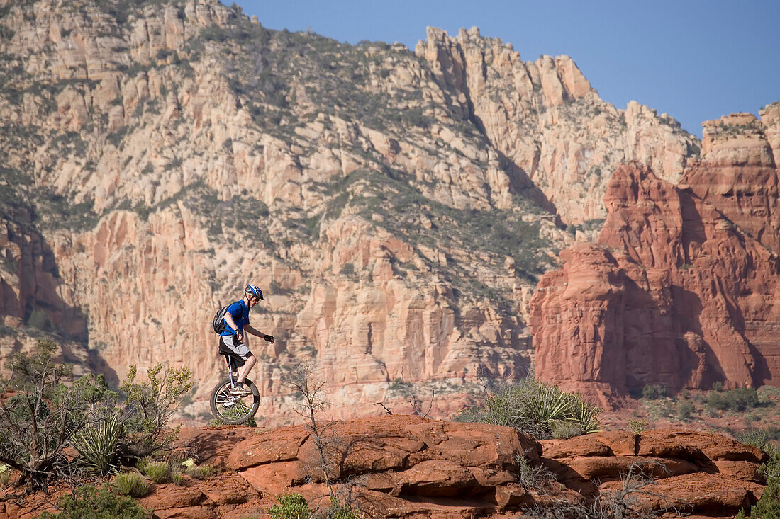 A man on a unicycle rides along a rocky ridge in the Arizona desert.; Sedona, Arizona.