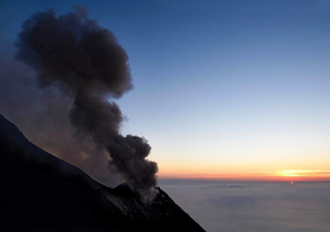 Smoke and ash rise up out of a volcano crater and into the evening sky.; Stromboli Island, Italy.