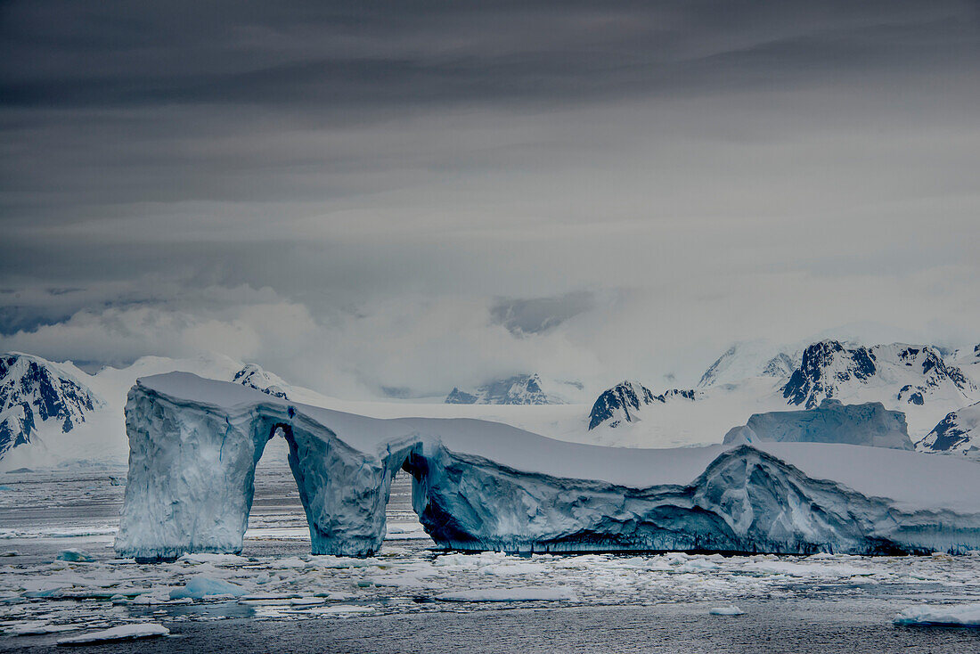 Iceberg with arches and snow covered mountains in the Penola Strait, a point at the far southern end of most Antarctic cruises; Antarctica