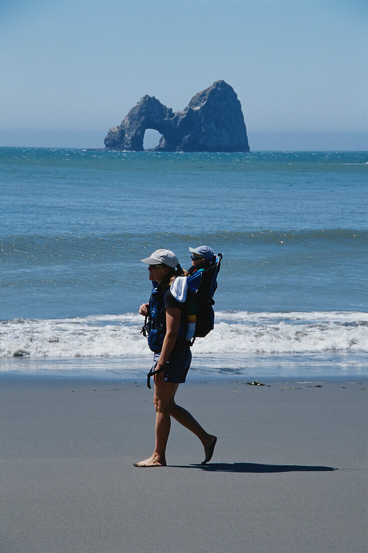 Frau und Baby im Rucksack wandern am Strand entlang; Arch Rocks, Oregon.