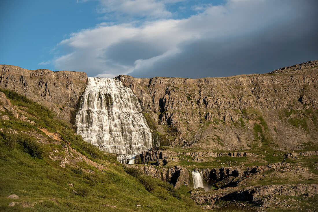 Dynjandi Waterfall with a dark cloud formation above the sunlit cliffs, is the the main waterfall of Dynjandi, a series of 7 waterfalls on the Arnarfjörður and the largest waterfalls in the Westfjords of Iceland; Arnarfjordur, Westfjords, Iceland