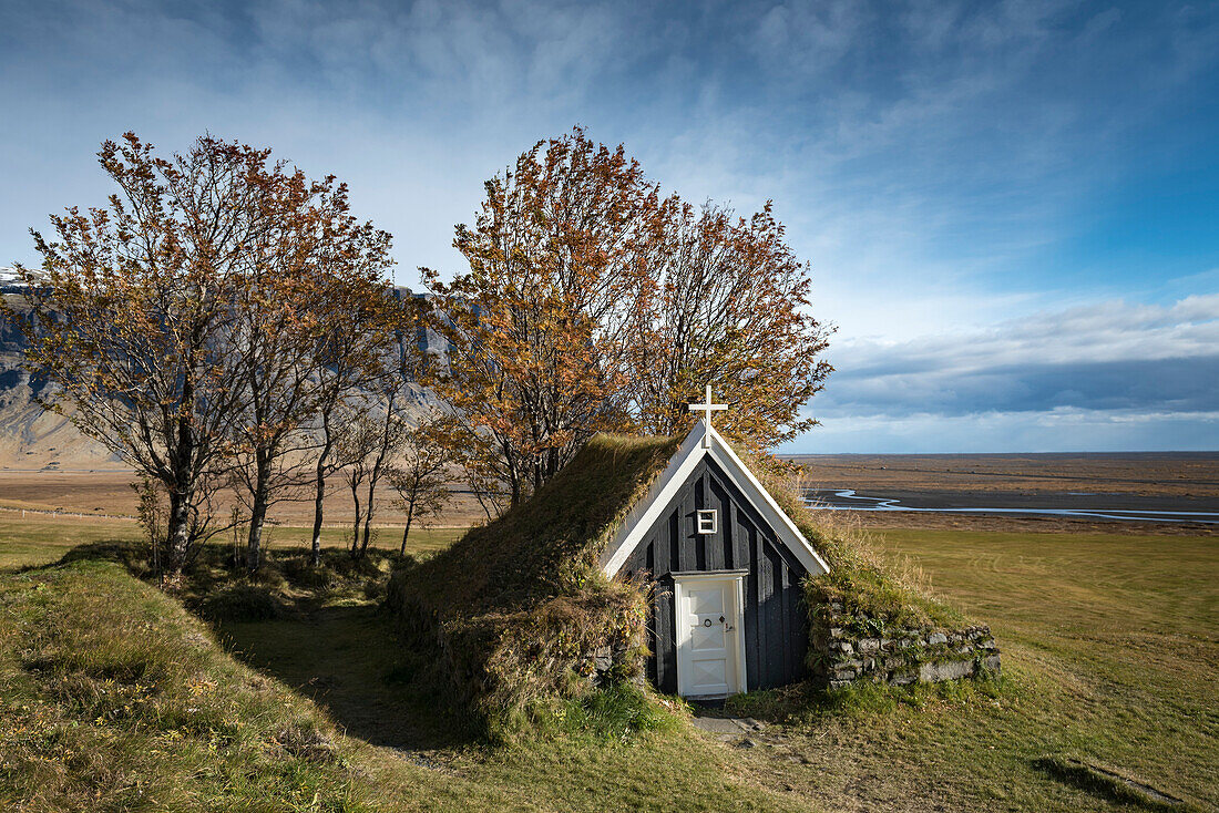 Kleine, schwarz gestrichene Kirche mit Grasdach zwischen Bäumen in Herbstfarben; Island