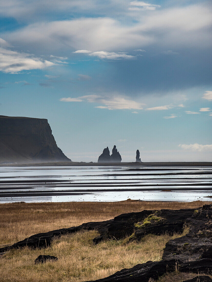 Blick auf Klippen und schwarzen Sandstrand, Reynisfjara Beach, an der Südküste Islands, nahe der Stadt Vik, mit schimmerndem Wasser im Vordergrund; Island