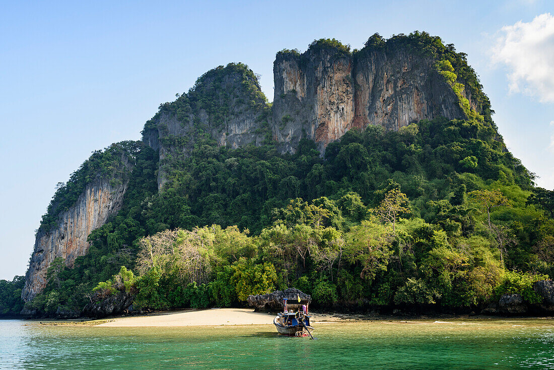 Blick auf einen kleinen Strand entlang der Küste einer tropischen Insel mit Klippen aus Karstfelsen und einem thailändischen Longtailboot, das am Ufer vertäut ist; Phang Nga Bay, Thailand