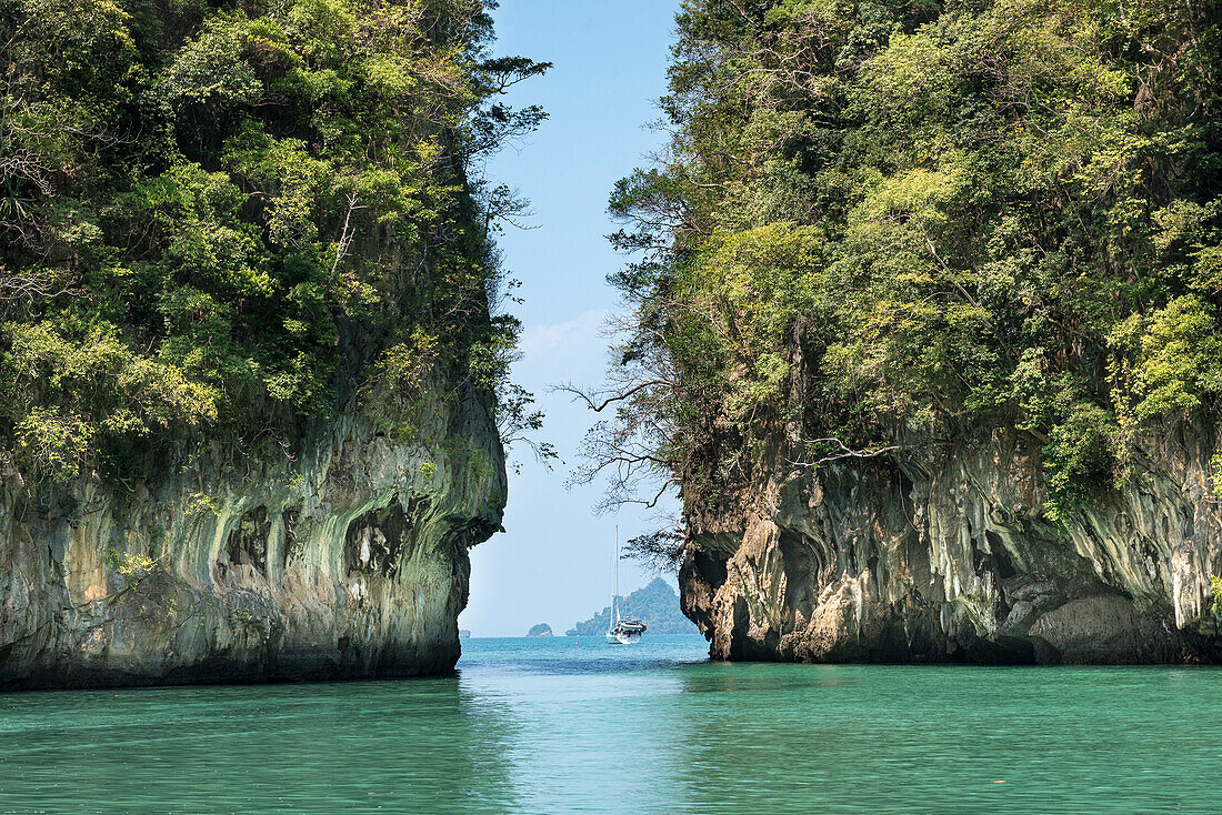 View of a yacht anchored in the green, turquoise colored waters in between too vast, karst rock formations creating a tranquil scene; Phang Nga Bay, Thailand