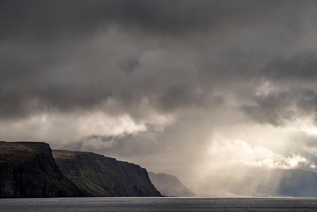 Die isländische Küstenlinie von Latrabjarg, als sich die dichten Wolken für die Sonnenstrahlen lösen. Latrabjarg ist die größte Meeresklippe Islands und der westlichste Punkt Europas und beherbergt die größte Konzentration von Seevögeln in Island; Latrabjarg, Island