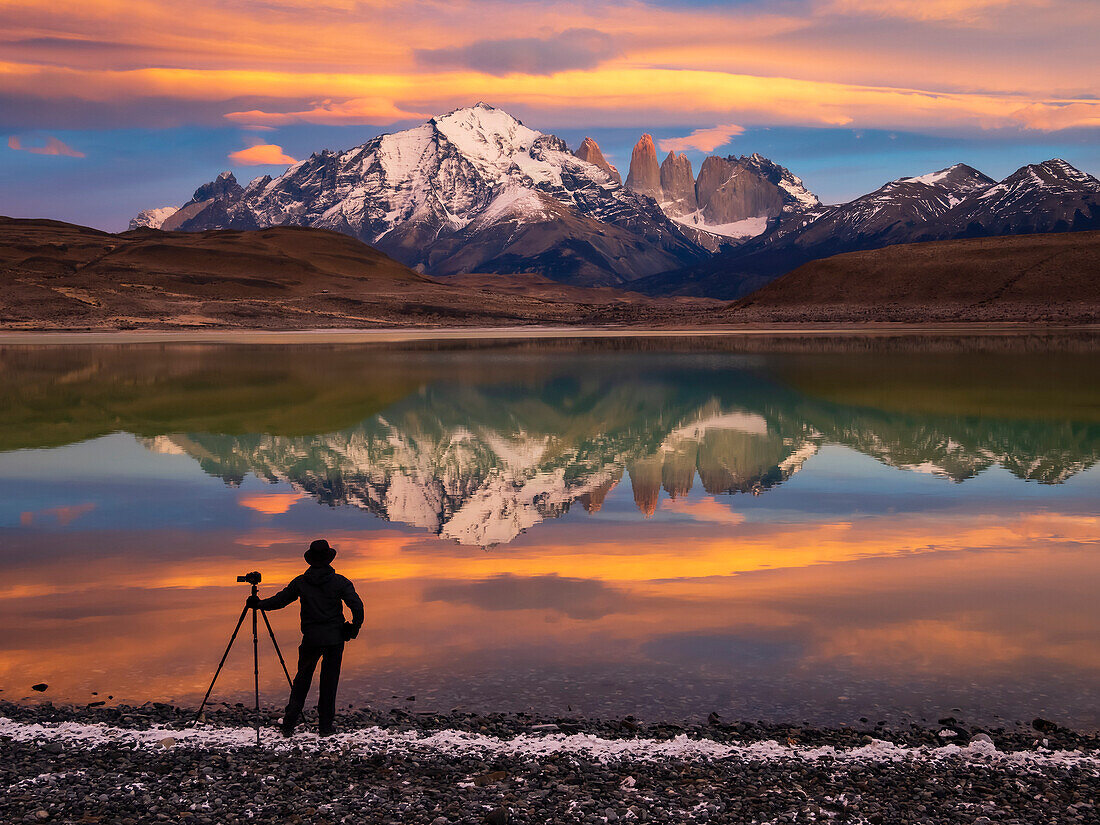 View taken from behind of a silhouette of a photographer with a camera tripod standing at the water's edge at Lago Azul at sunrise tracking pumas; Torres del Paine National Park, Patagonia, Chile