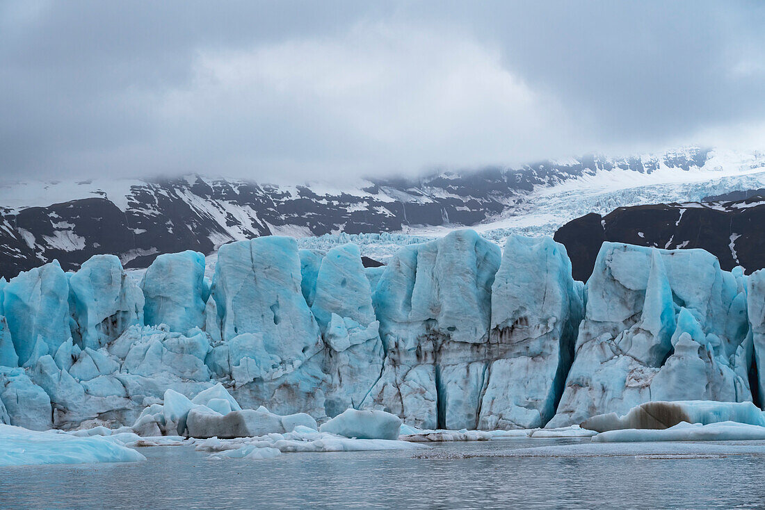 Views of the blue ice of the Fjallsjokull Glacier stands out against the gray, misty clouds over the snow-covered mountains viewed from the Fjallsarlon Glacier Lagoon, at the south end of the famous Icelandic glacier Vatnajökull in Southern Iceland; South Iceland, Iceland