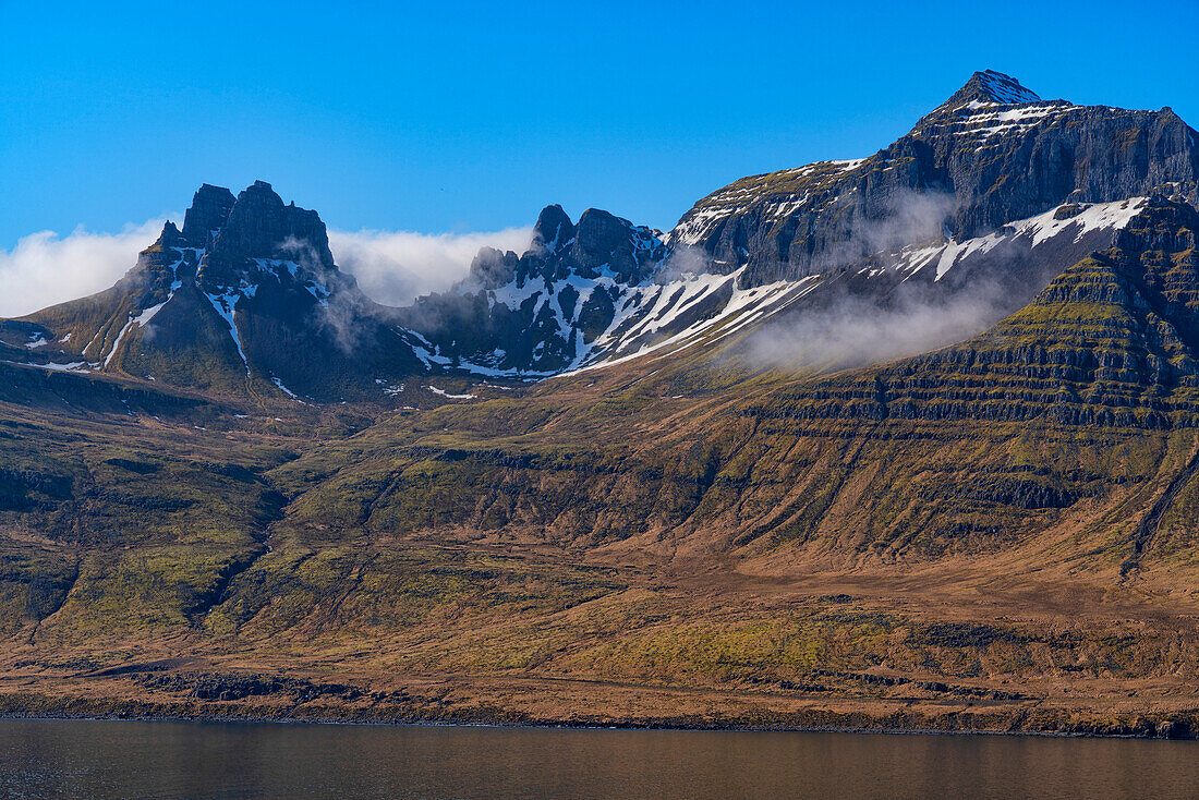 Nahaufnahme des gebirgigen Geländes der Ostfjorde mit blauem Himmel und nebligen Bergwolken, die eine atemberaubende Landschaft zum Durchreisen schaffen; Ostisland, Island