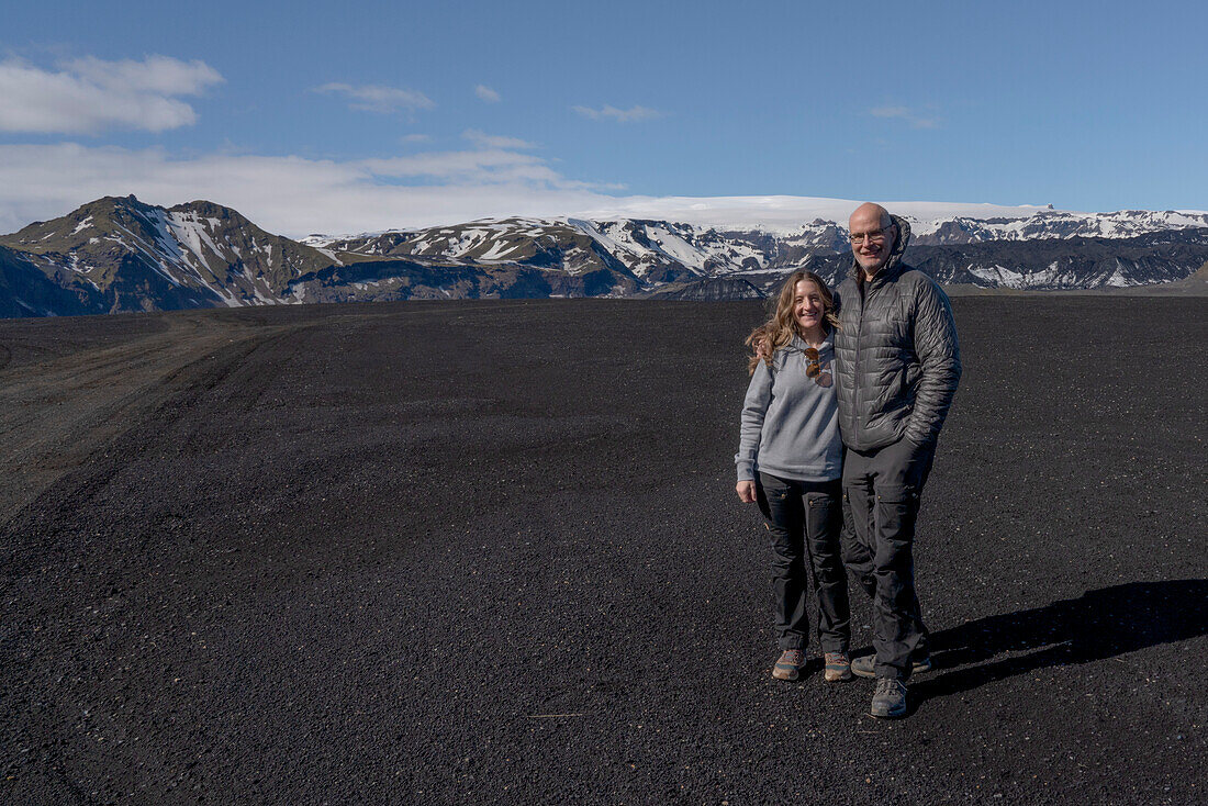 Close-up view of a smiling couple posing for the camera, standing in front of a massive landscape of black sand with a mountain ridge and a blue sky in the background; Iceland