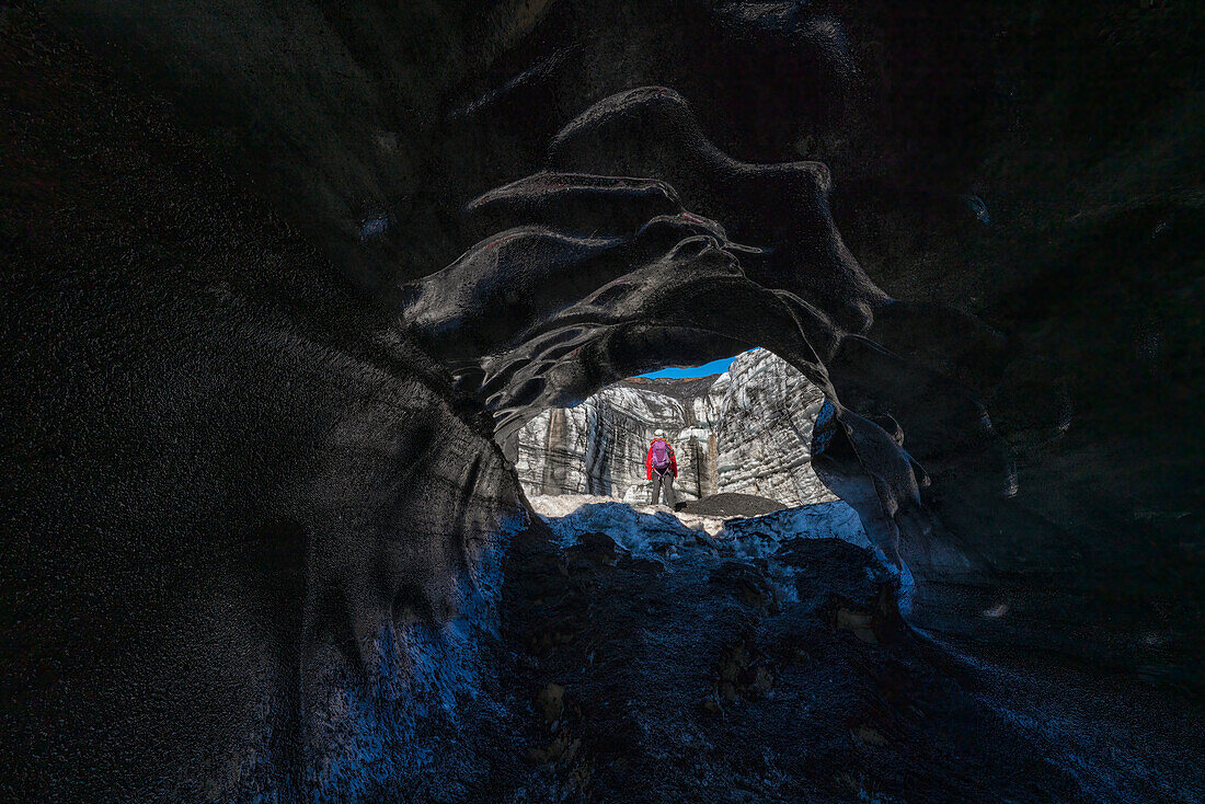Blick durch eine Eishöhle auf eine Frau, die einen Gletscher erkundet, während sie in Island unterwegs ist; Vik, Südisland, Island