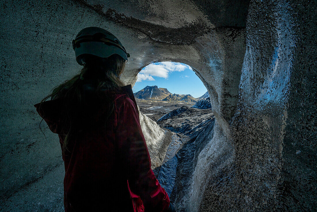 View through an ice cave of a woman exploring a glacier while traveling in Iceland; Vik, South Iceland, Iceland