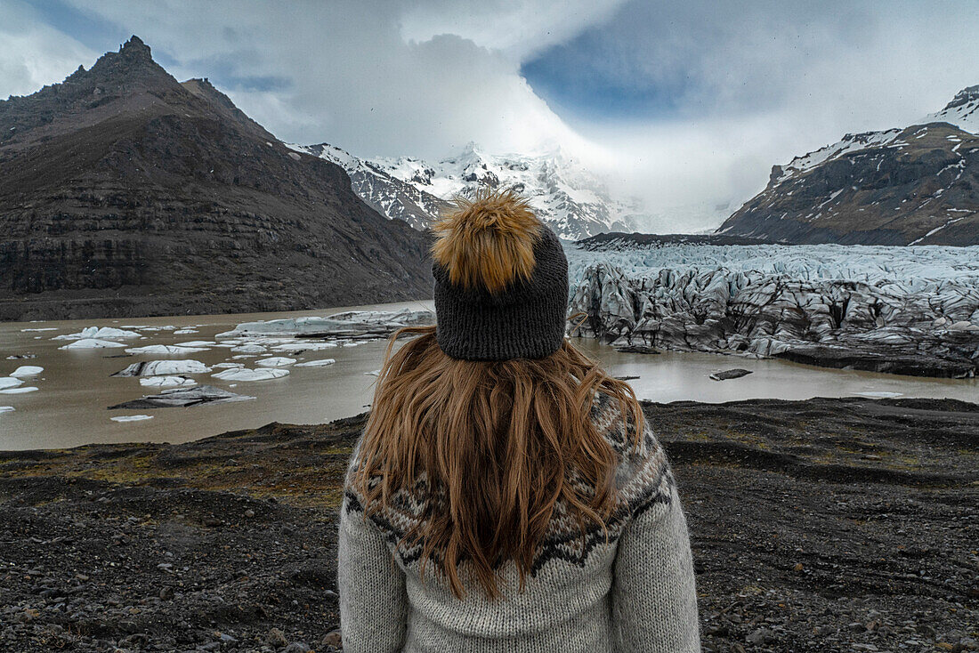 Close-up view of a woman with blond hair, taken from behind, wearing a woolen hat and an Icelandic sweater overlooking a glacier and lagoon on the South Coast; South Iceland, Iceland