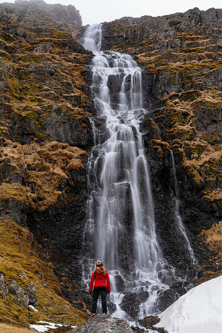 Blick von hinten auf eine Frau, die vor einem Wasserfall an der Strandir-Küste in der Nähe der Stadt Djupavik im Nordwesten Islands steht; Djupavik, Westfjorde, Island
