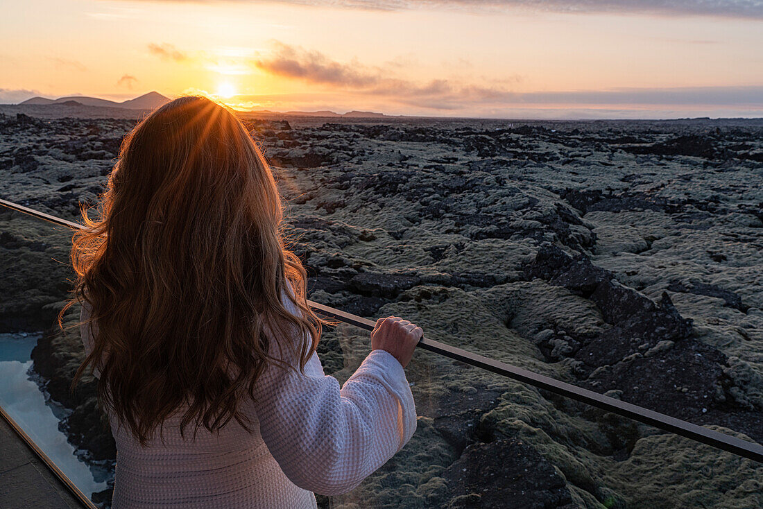 Frau steht im Bademantel auf einem Balkon mit Blick auf die Lavafelder in Südisland; Blaue Lagune, Südisland, Island