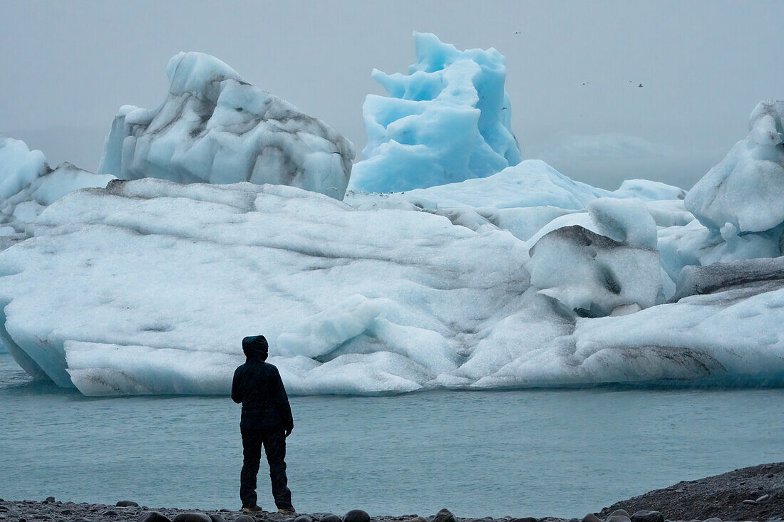 Silhouette einer Frau im Schneeanzug, die am Ufer steht und die wunderschönen Eisberge und die erstaunlichen, blauen Eisformationen und Formen der J?s???lacier Lagoon am südlichen Ende des berühmten isländischen Gletschers Vatnaj?l in Südisland betrachtet; Südisland, Island
