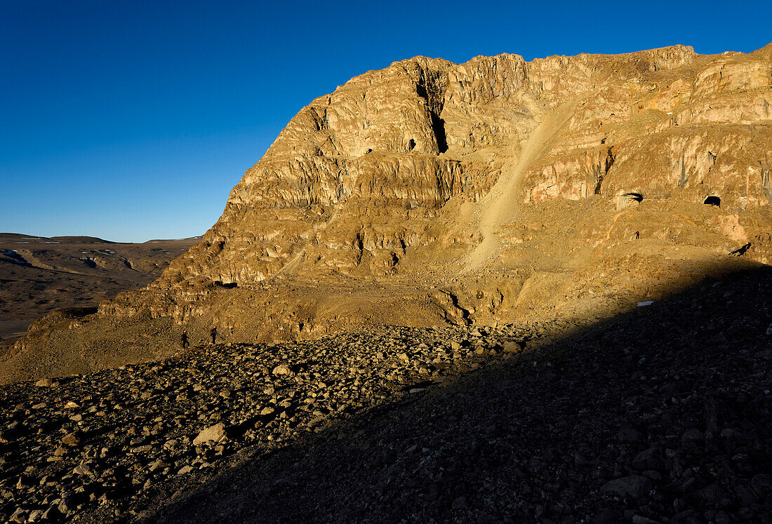 As the evening sunlight arrives, two expedition team members walk along a ridge bathed in sunlight towards another team member standing upon a large lump of rock. In the distance, a team member stands in blue in the middle of the righthand entrance to The U-shaped cave. The giant scree slope, the largest of all the scree slopes of rocks the team endured on this expedition drops away in the middle of the cliff face, providing the access they needed to get into The Crystal Palace