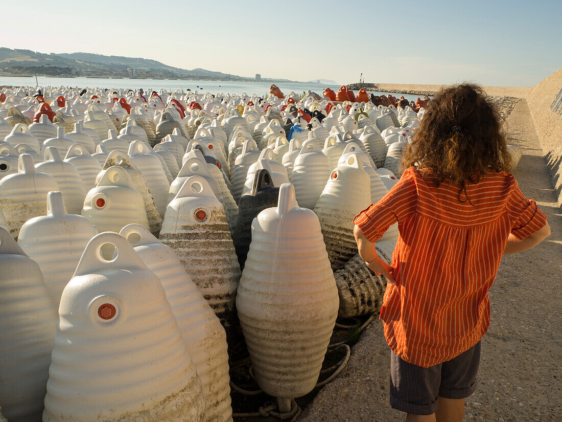 Große Menge an Schwimmern für Muschelkulturen am Ufer mit Hügeln im Hintergrund und Blick von hinten auf eine Frau, die im späten Nachmittagslicht auf das Meer hinausschaut; Porto San Giorgio, Marken, Italien