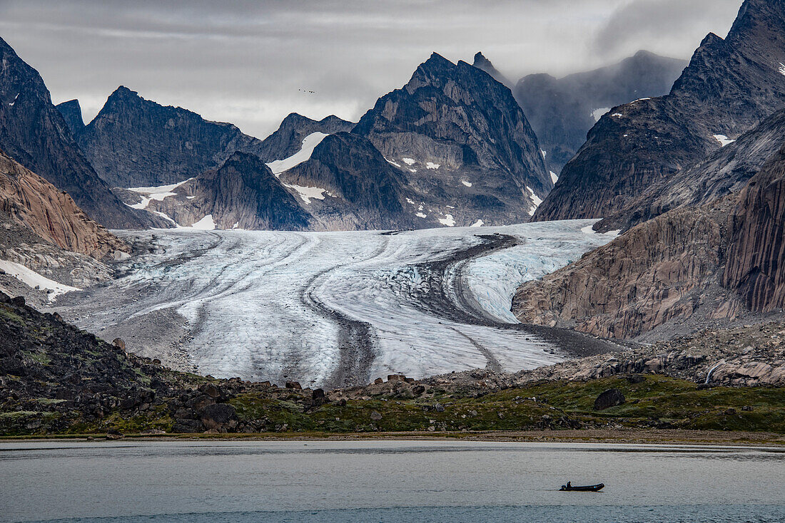 View of glacier flowing through the mountain peaks at the Southern tip of Greenland with silhouette of a boat passing by in the grey waters of Prins Christian Sund; Southern Greenland, Greenland