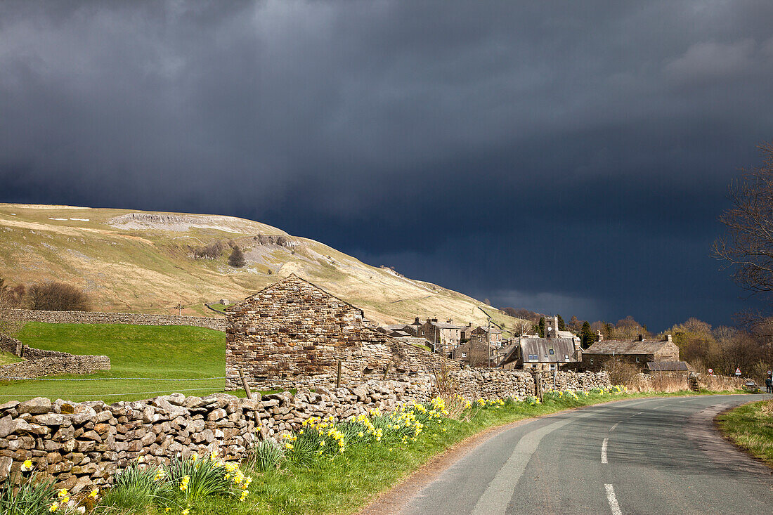 Eine Steinmauer und Gebäude entlang einer gepflasterten Straße mit dunklen Gewitterwolken über dem Kopf; Muker Dorf Yorkshire England