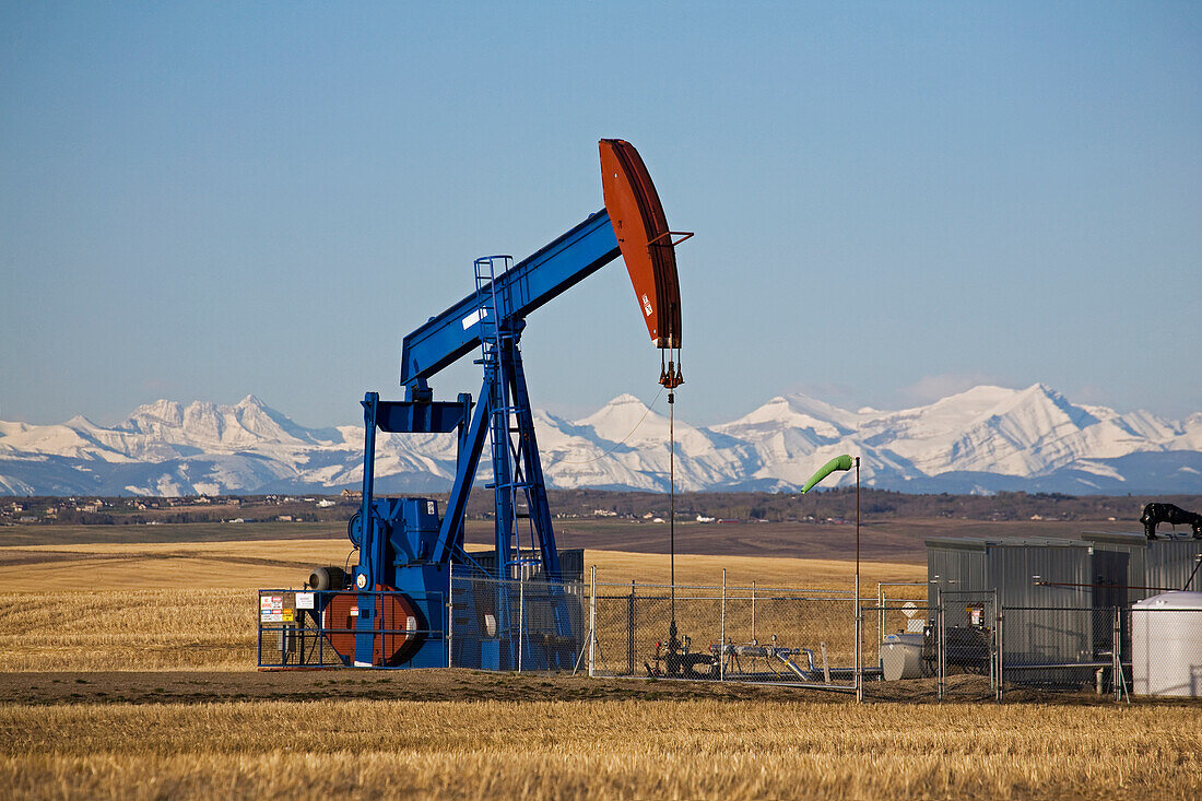 Pumpjack auf einem Feld mit schneebedeckten Bergen und blauem Himmel im Hintergrund; Alberta, Kanada