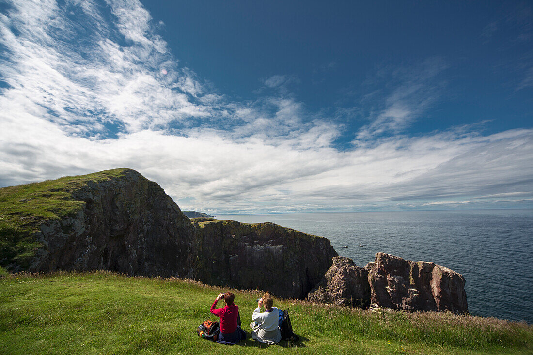 Two People Sitting On The Grass Looking Through Binoculars At The Rocky Promontory At St. Abb's Head; Scottish Borders Scotland