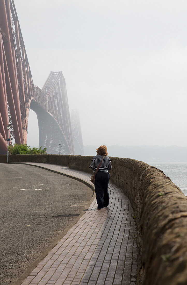 A Woman Walking On A Pathway On The Water's Edge Towards Forth Bridge; North Queensferry Scotland
