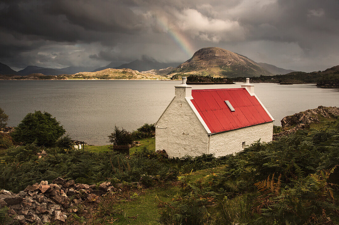 Ein Cottage mit rotem Dach am Wasser mit einem Regenbogen in der Ferne; Applecross Peninsula Highlands Schottland