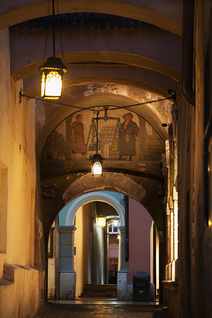 Arched passageway with fresco and street lamps at night; Bolzano, Alto Adige, Italy