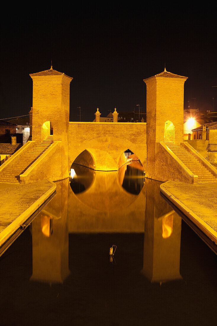 Italy, Emilia-Romagna, Comacchio, Stone bridge with towers and steps reflecting in water, night