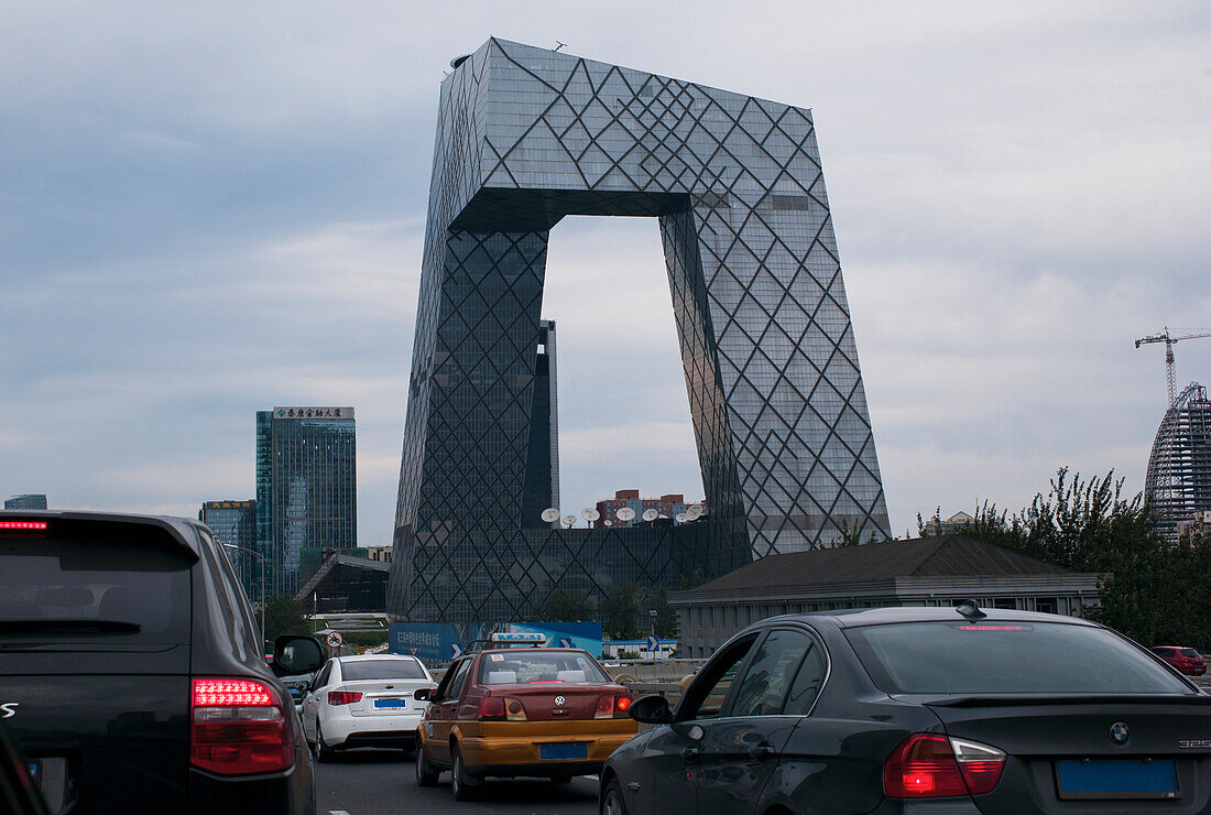 Autos im Verkehr auf der Straße mit einem einzigartig geformten modernen Gebäude im Hintergrund; Peking, China