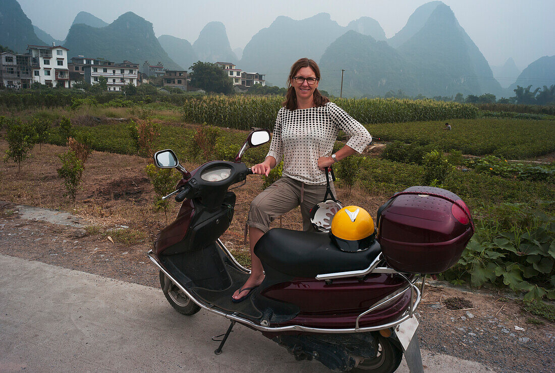 A Woman Poses With Her Motor Scooter With The Peaked Mountains In The Background; Yangshuo, China