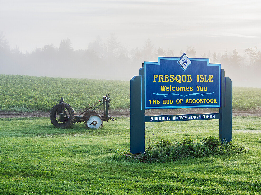 Welcome Sign To Presque Isle, Maine On A Foggy Summer Morning With Farm Equipment, A Potato Field And Dewy Grass Surrounding It; Presque Isle, Maine, United States Of America